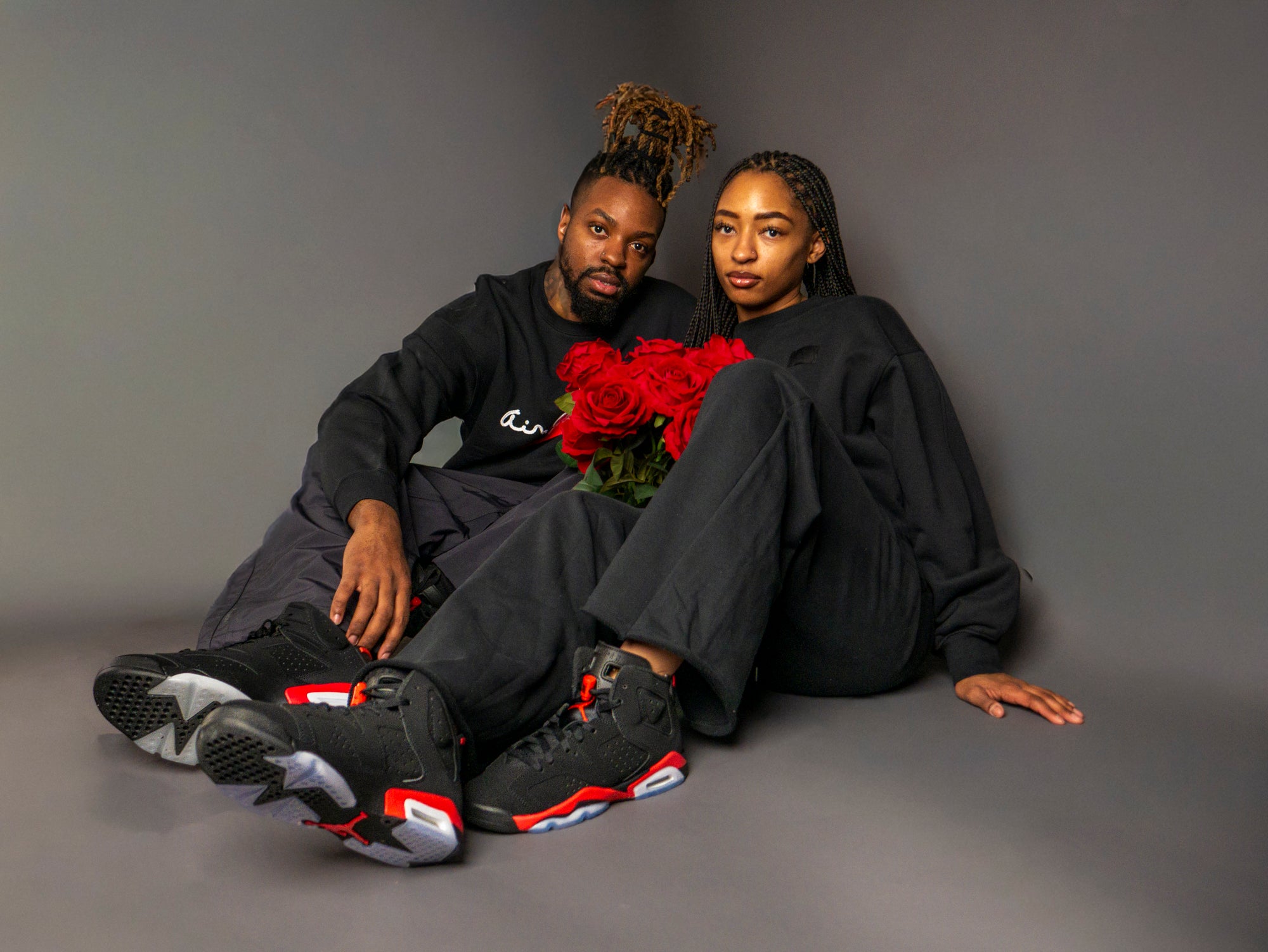 Two people sitting on a gray floor with a bouquet of red roses, wearing matching black Air Jordan outfits and Air Jordan 6 Retro "Infrared Salesman" sneakers.