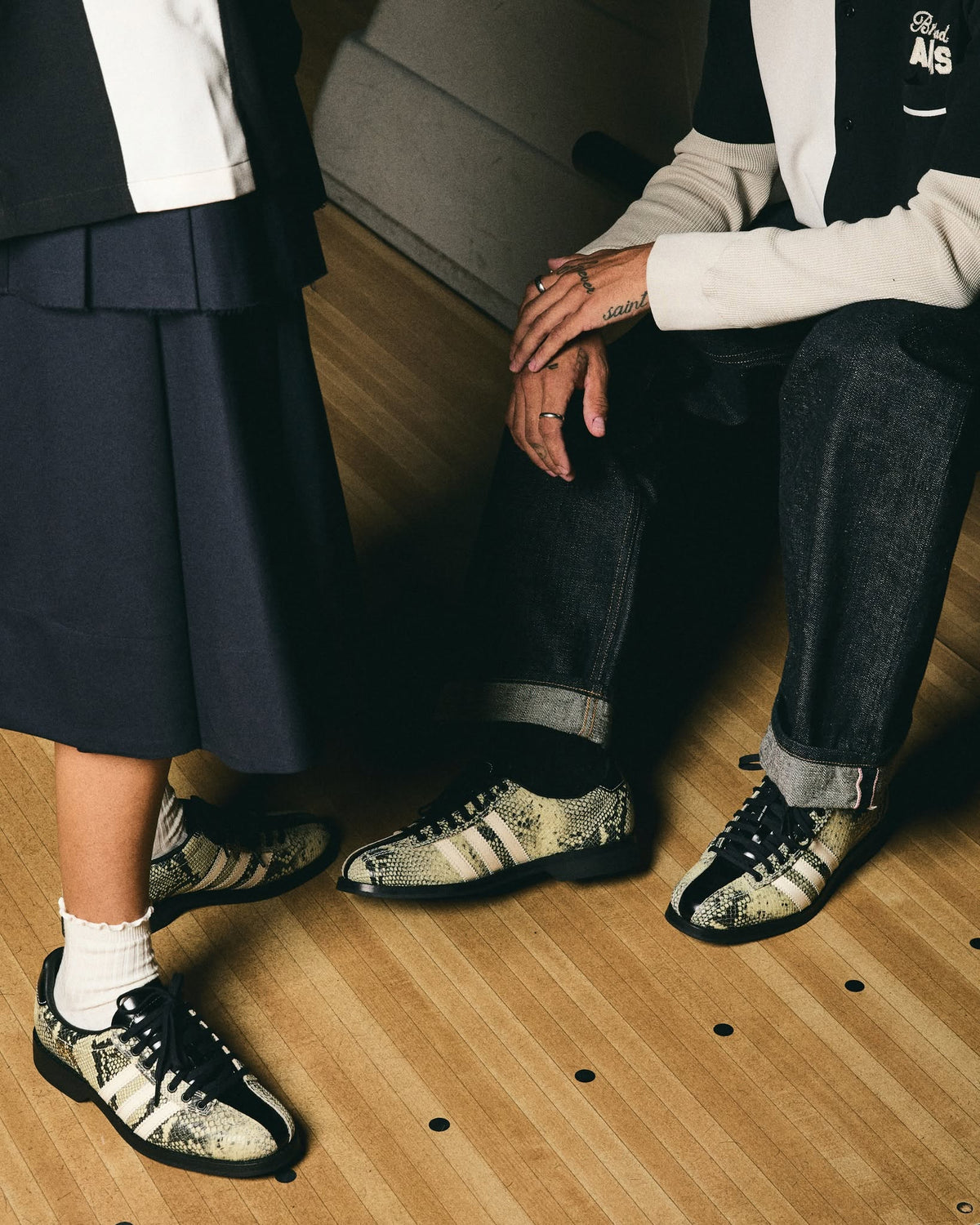 Two models wearing matching black, white, and gray snakeskin-patterned sneakers, standing and sitting on a light wood floor.